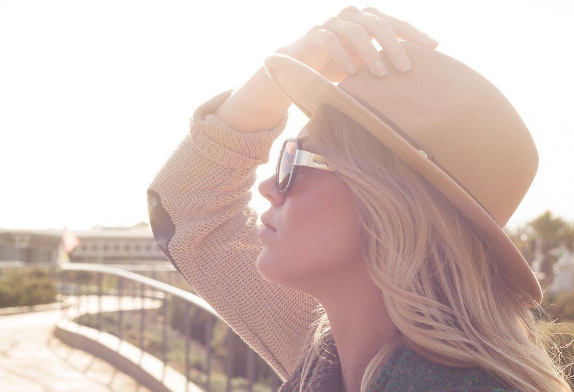 Woman in sunglasses and hat, looking up, holding hat with sunlight in the background.