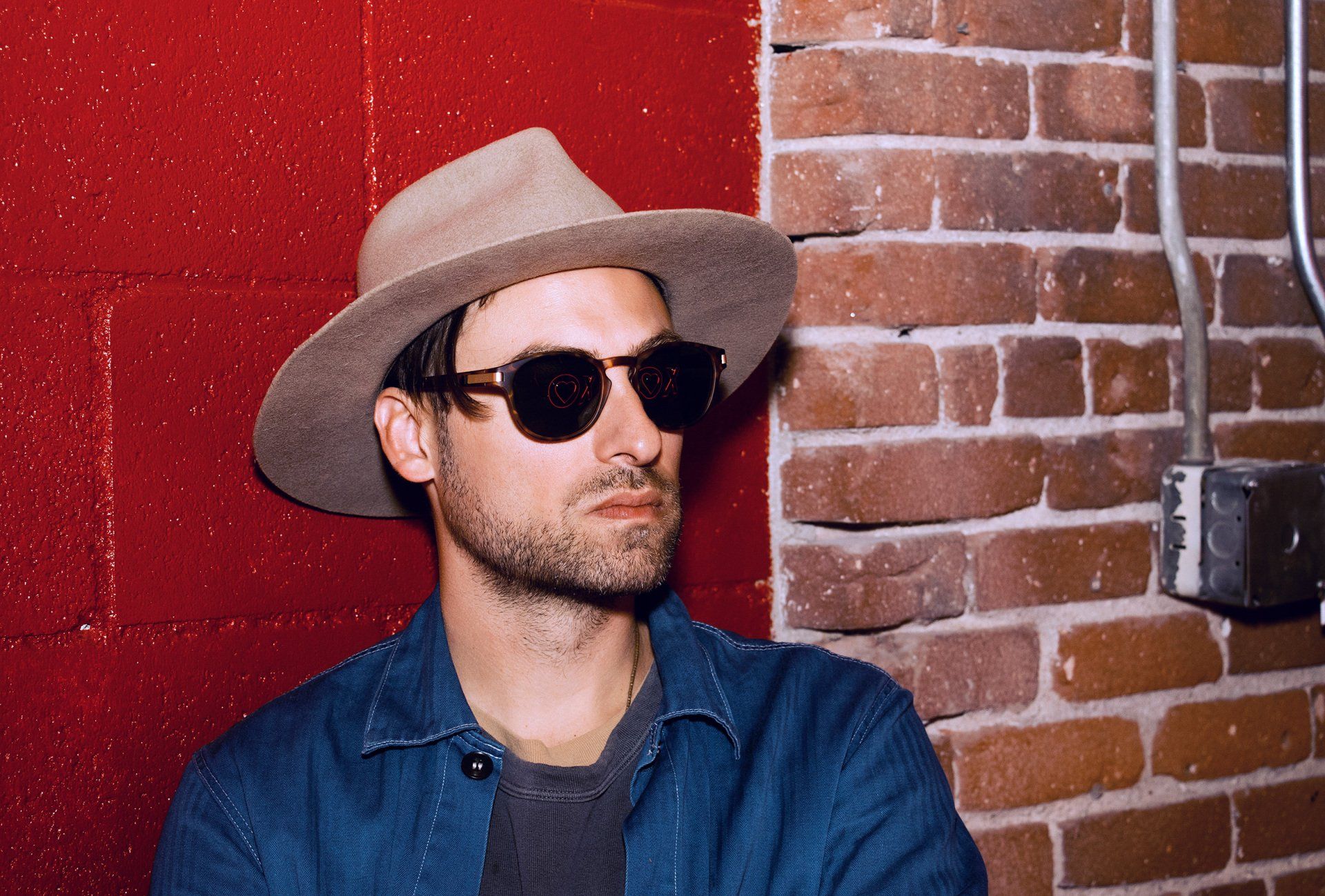 Man in fedora and sunglasses leans against brick and red wall.