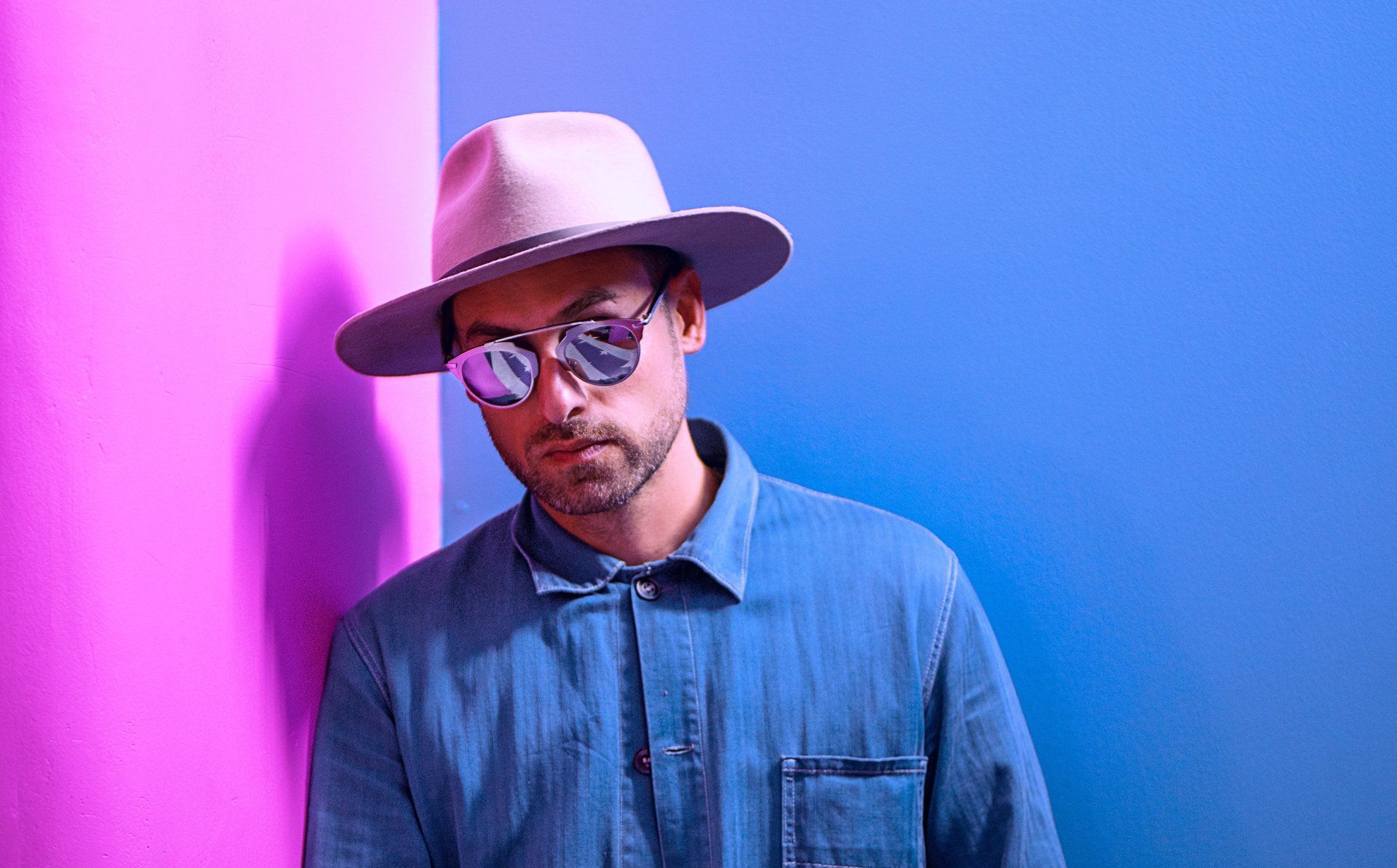 Man wearing sunglasses and a hat, leaning against a pink and blue wall.