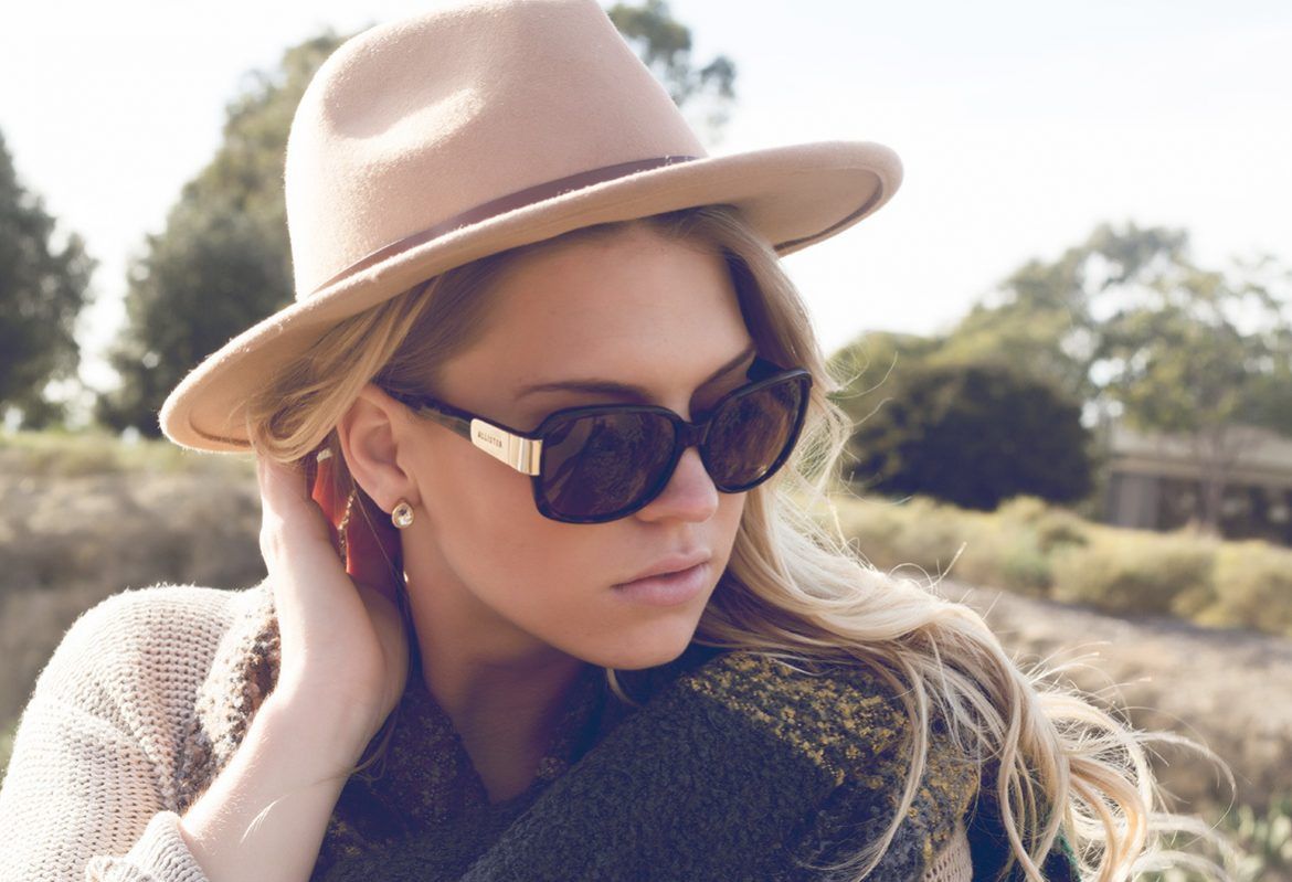 Woman in hat and sunglasses outdoors, touching her ear.