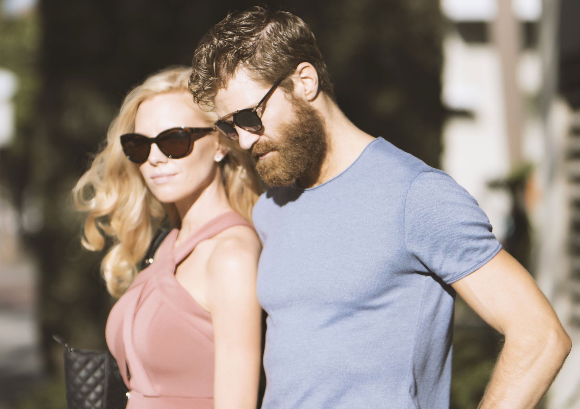 A couple wearing sunglasses walk outside in the sunlight. The woman wears a pink dress and the man a blue shirt.