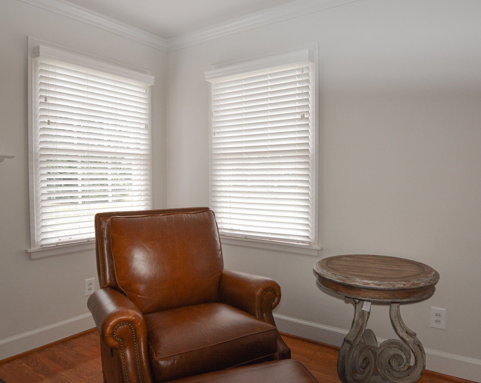 A living room with a brown leather chair , ottoman , table and two windows.