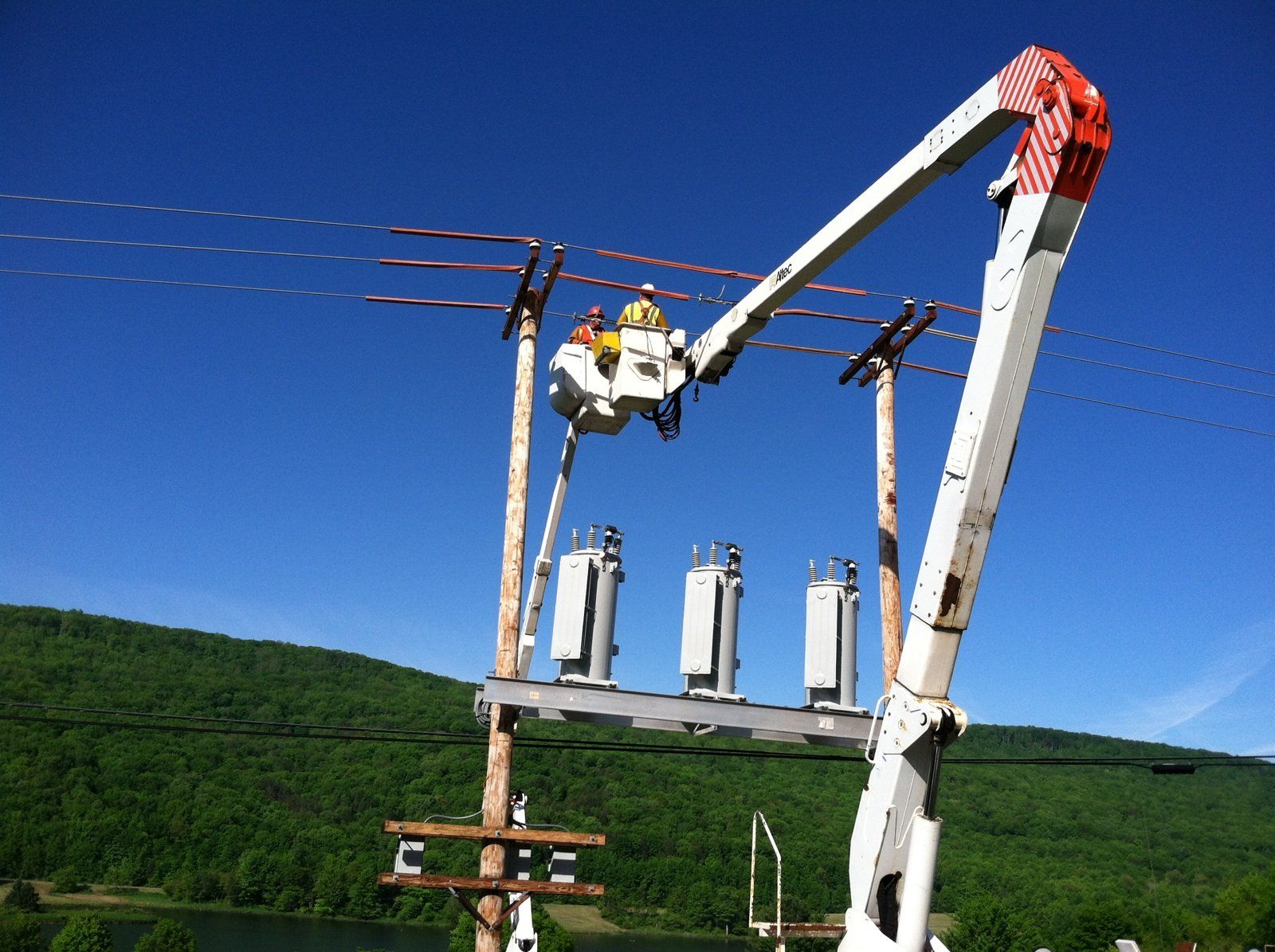 Linemen working on power lines using a bucket truck against a blue sky, near a lake and trees.