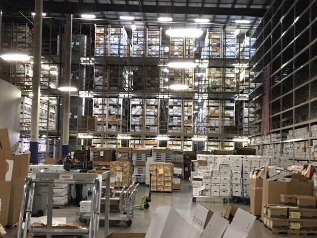 Warehouse interior with tall shelving packed with boxes. Cardboard boxes and carts are in the foreground.