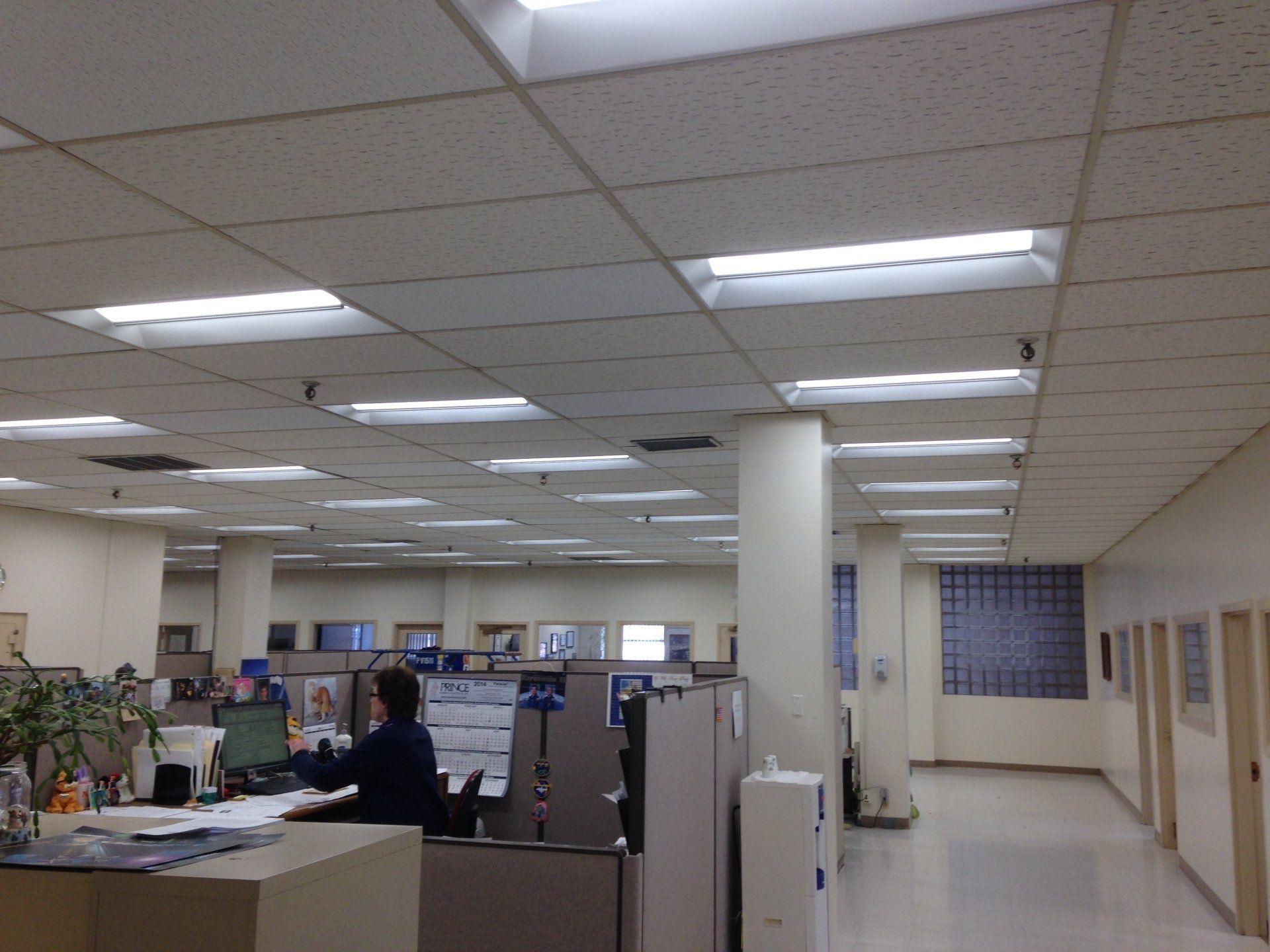 Office interior with fluorescent lights and cubicles. A person sits at a desk, windows and doors along a wall.