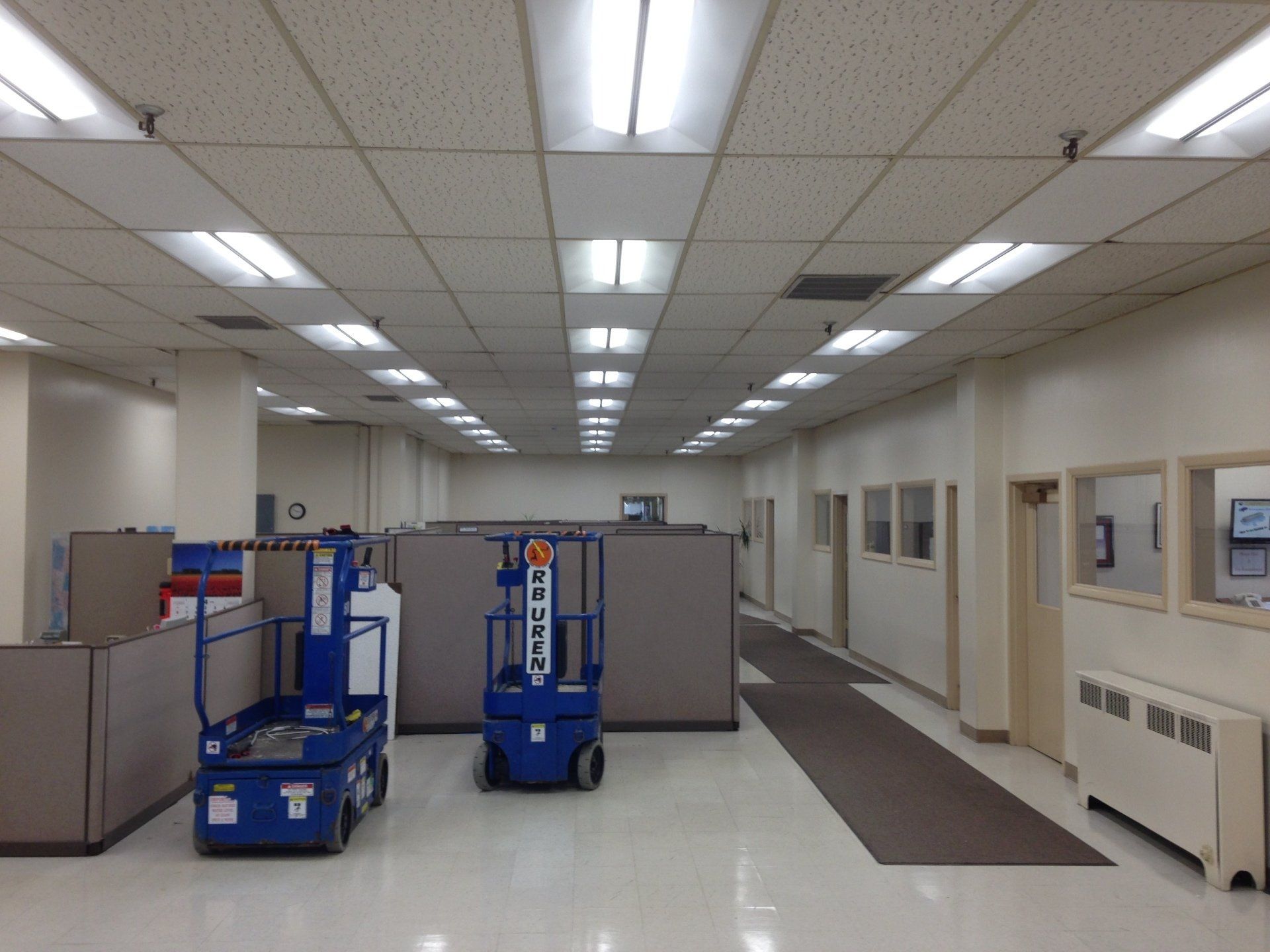 An empty office space with cubicles, two blue lifts, fluorescent lights, and a long hallway.