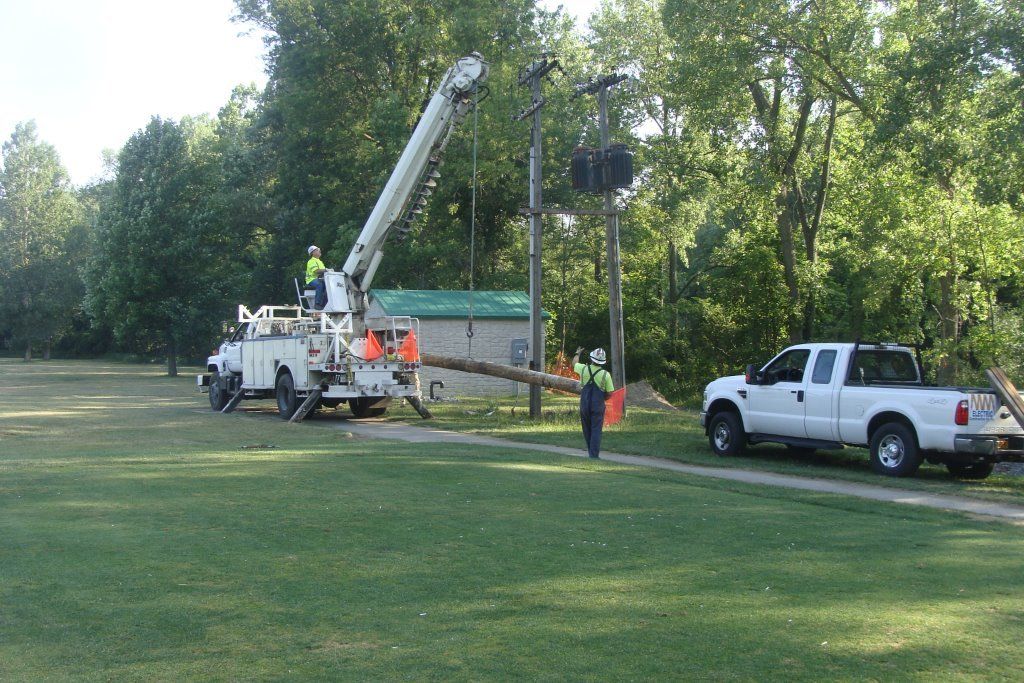 Workers in a bucket truck replacing a utility pole in a grassy park, next to a white pickup.