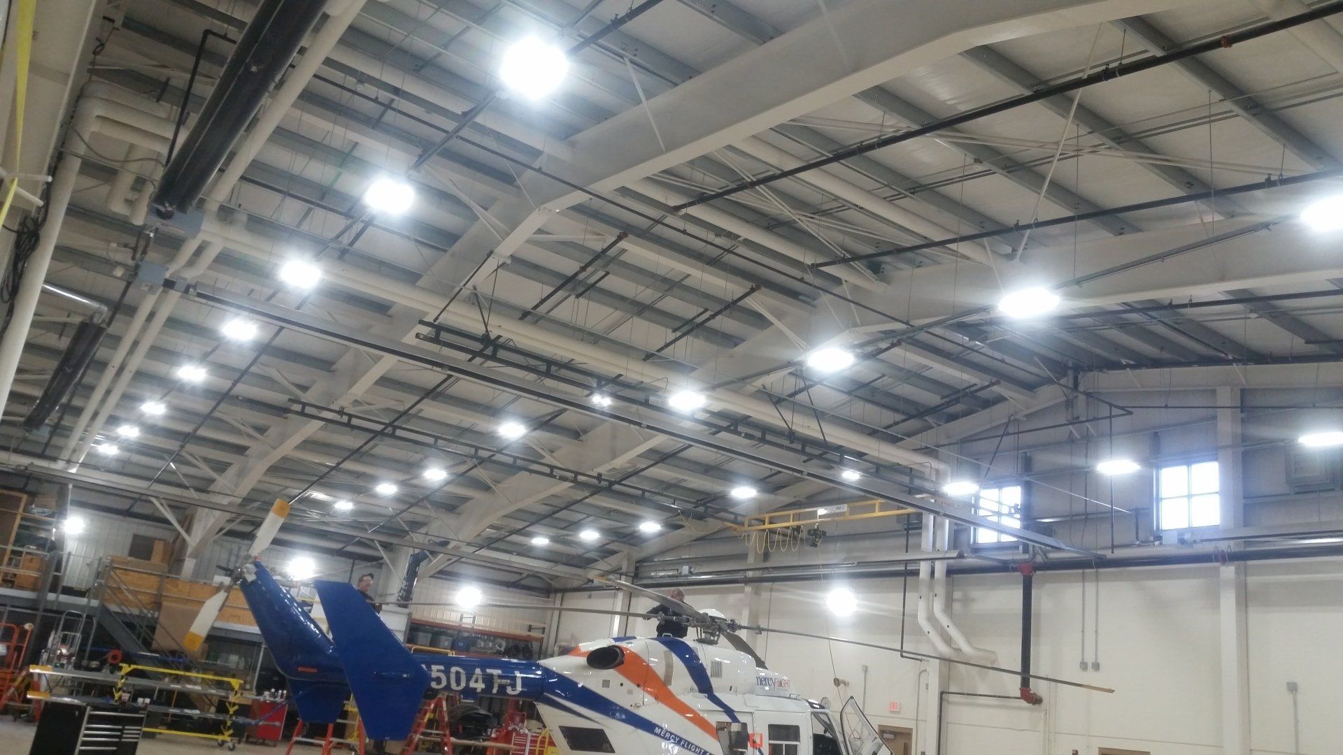 Helicopter inside a well-lit hangar with many overhead lights, metal roof, and equipment visible.