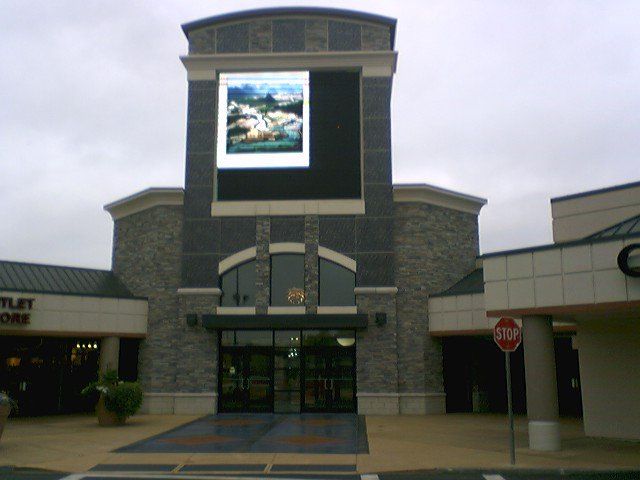 Shopping center entrance with large digital screen displaying artwork, cloudy sky.