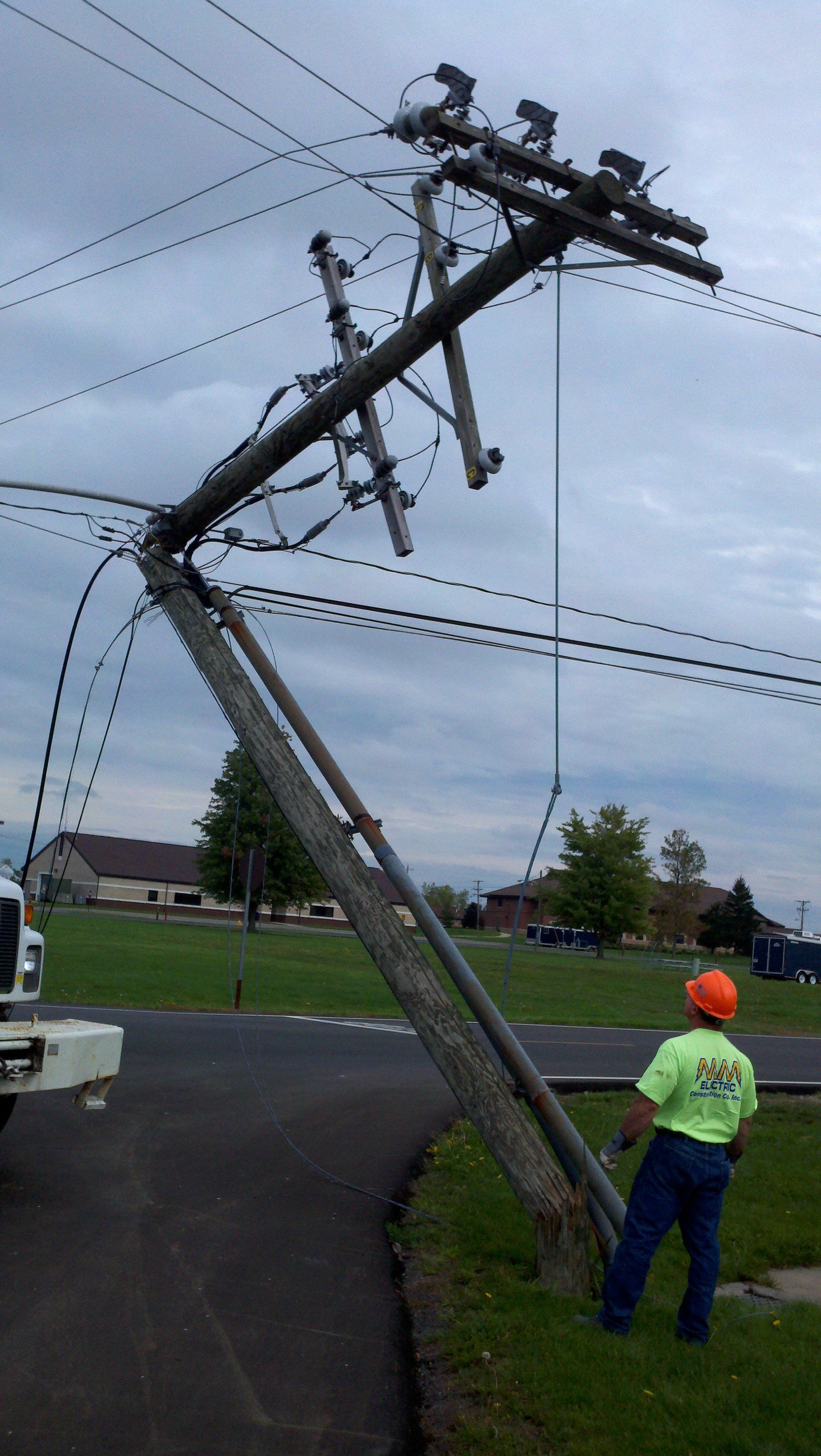 Leaning utility pole with attached wires; a worker in safety gear stands near it on a grassy area, alongside a road.