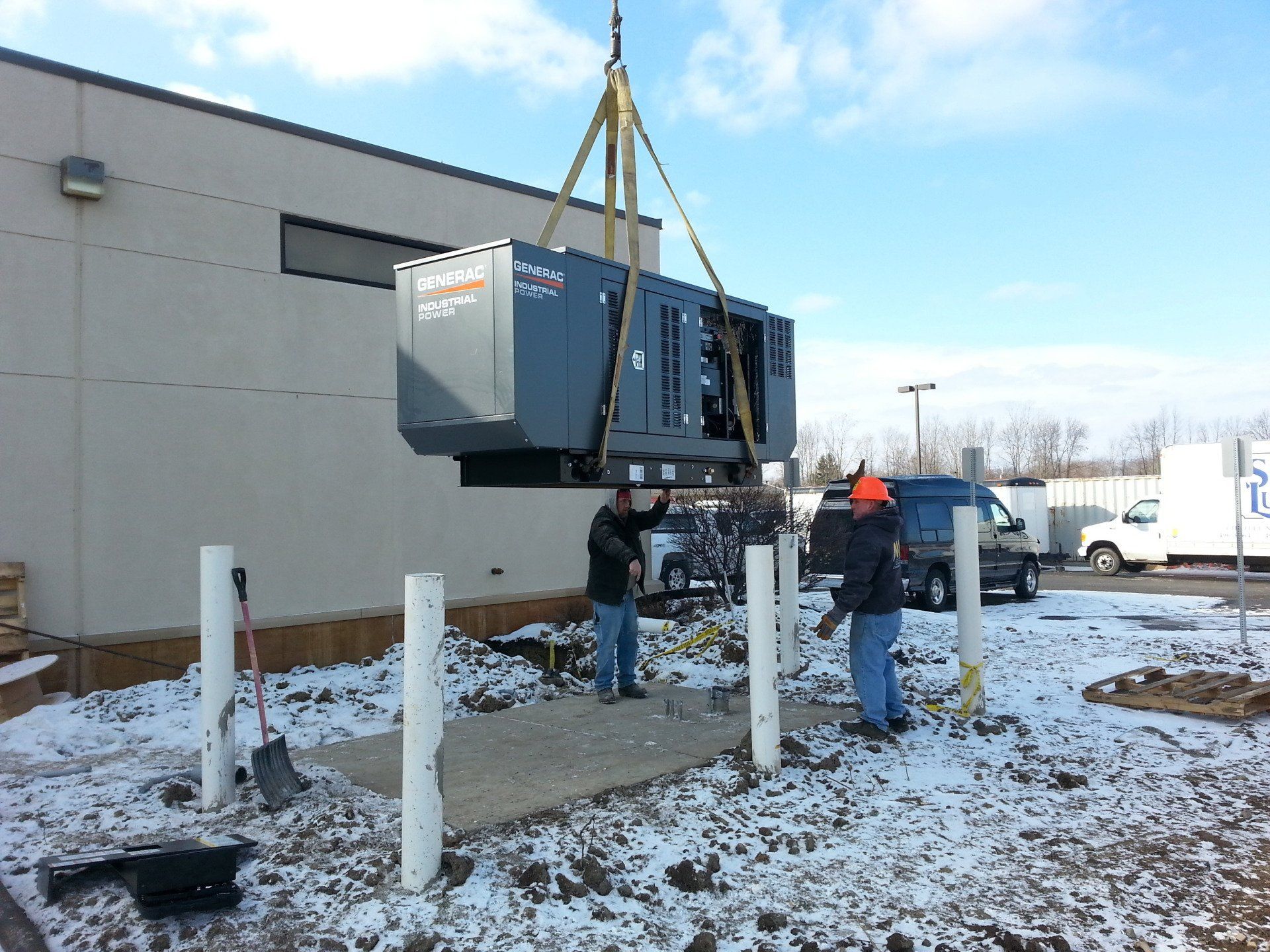 A commercial generator being lifted by a crane; two men below. Snowy outdoor setting.