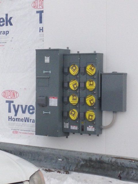 Gray electrical panel with multiple meter sockets mounted on a white wall with blue Tyvek wrap.