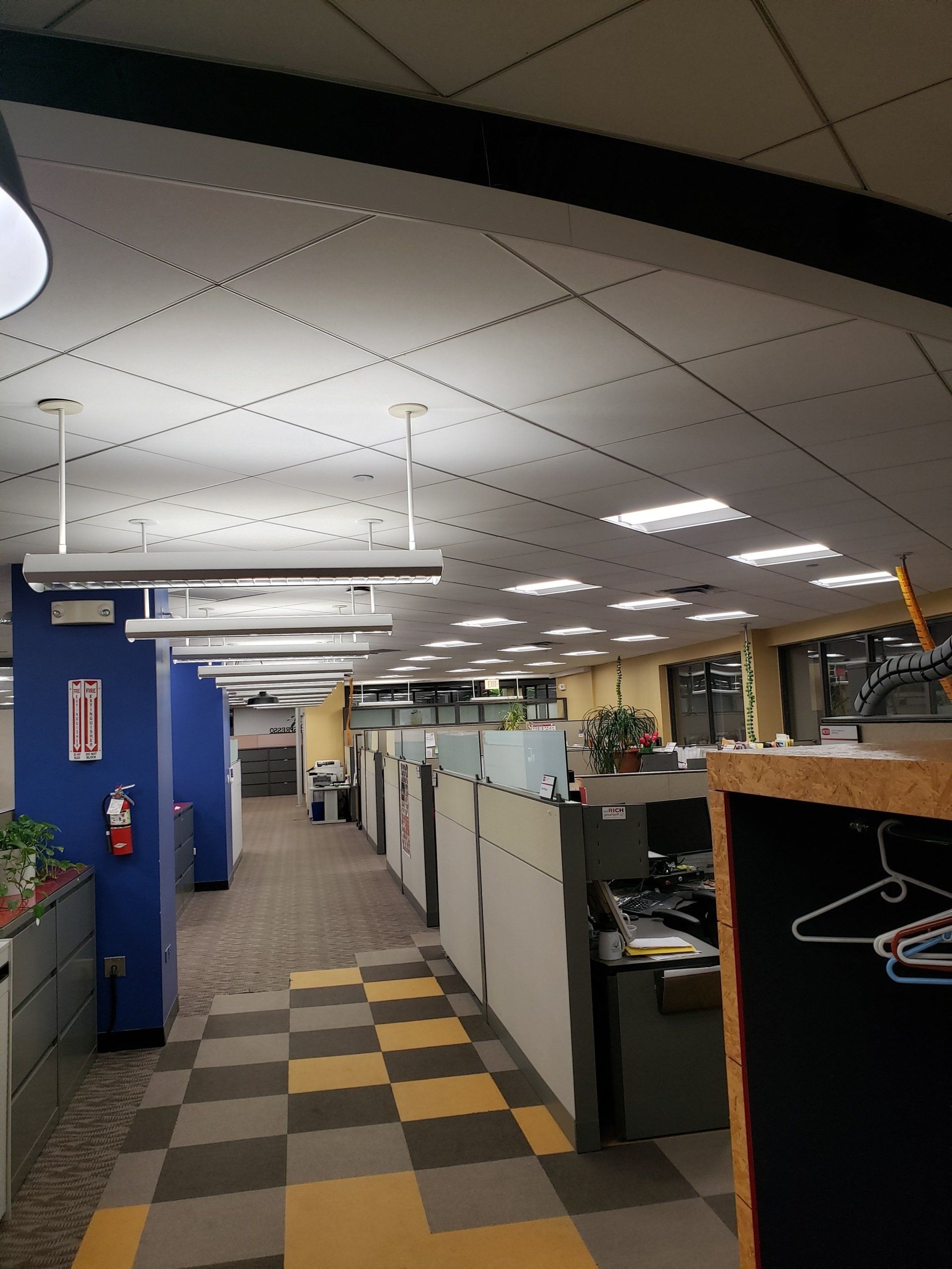Office with cubicles, blue and grey walls, grid ceiling, yellow and black checkered floor, fluorescent lights.