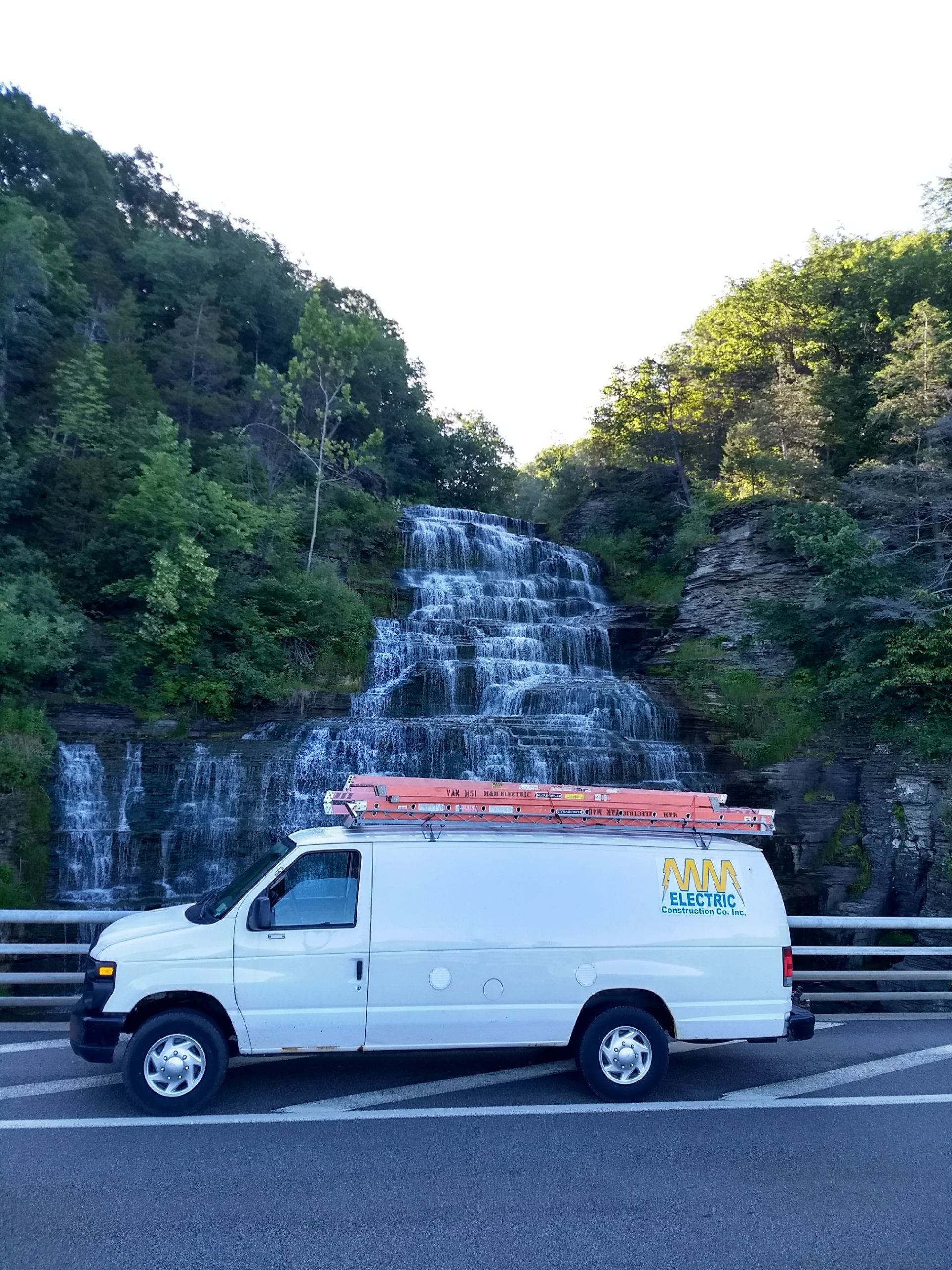 White service van parked on road in front of waterfall. Orange ladder on roof. Green trees surround.