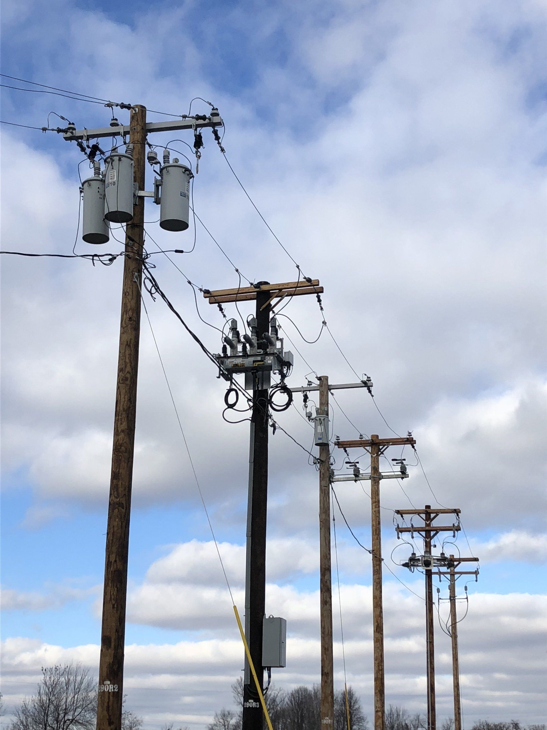 Power poles with transformers and wires against a cloudy blue sky.
