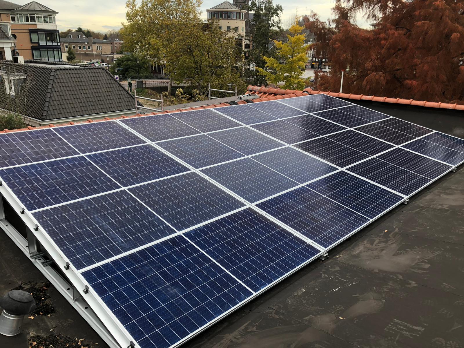 Zonnepanelen geïnstalleerd op een schuin dak bij schemering, met bomen en huizen op de achtergrond.