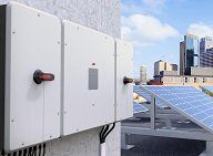 Witte elektrische schakelkasten gemonteerd op een dak naast zonnepanelen en met uitzicht op de skyline van de stad.