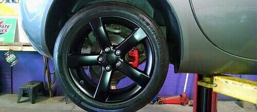 Black car rim with five spokes mounted on a vehicle in an auto repair shop with a red brake caliper visible.