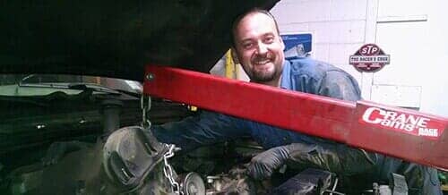 A smiling person in a blue shirt works on an engine inside a garage using a red hydraulic engine crane.