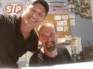 Two smiling men pose for a selfie in an office with a corkboard, an STP sign, and various photos on the blue wall.