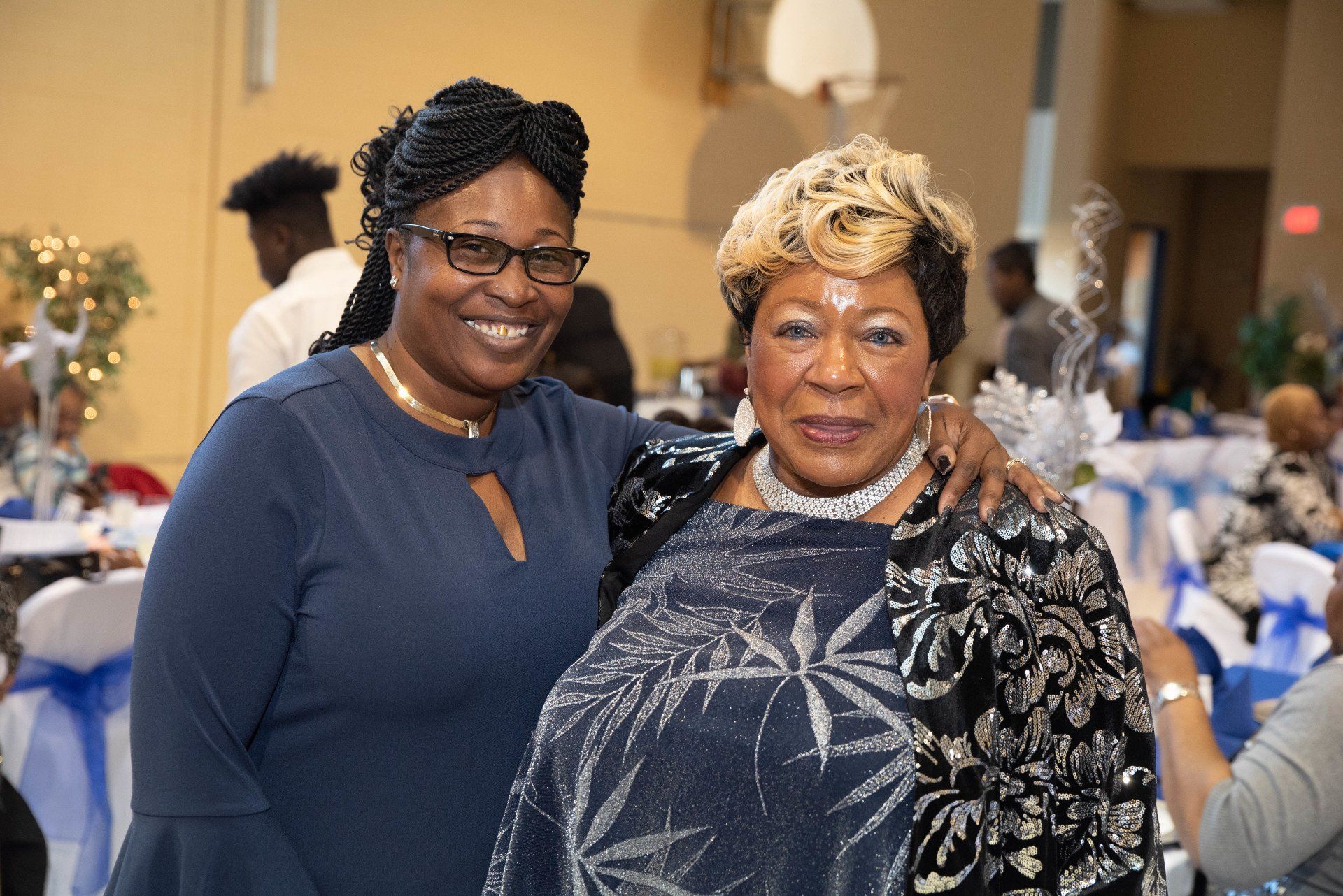 Two women smiling together at a decorated event. One wears glasses and a blue top; the other has blonde hair and a sparkly jacket.