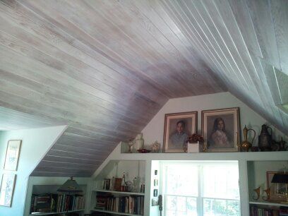 Sloping, wood-paneled ceiling painted pale gray. Portraits and bookshelves sit below, by a window.