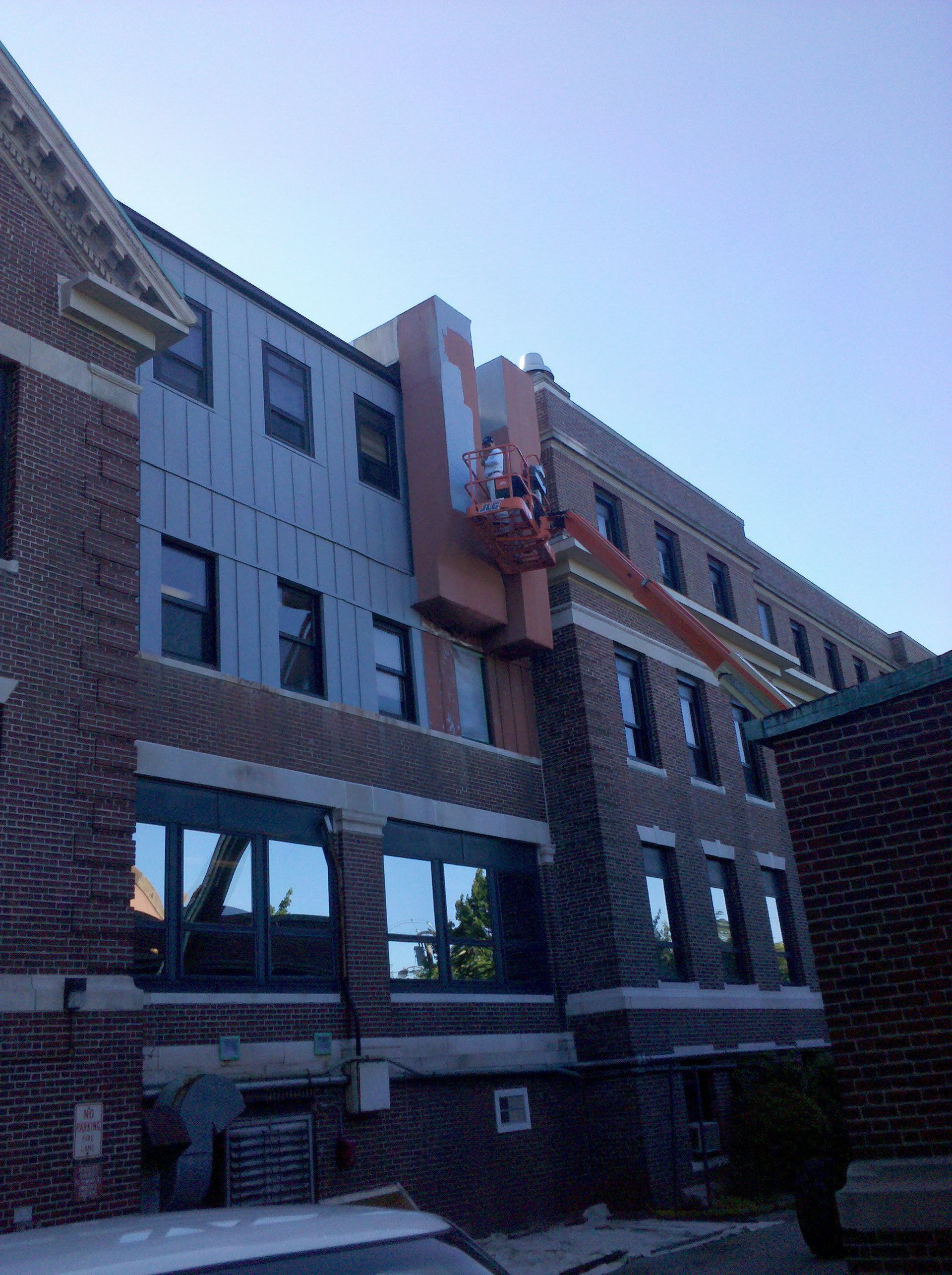 Construction on building, worker in orange lift. New light blue siding on section of brick building.