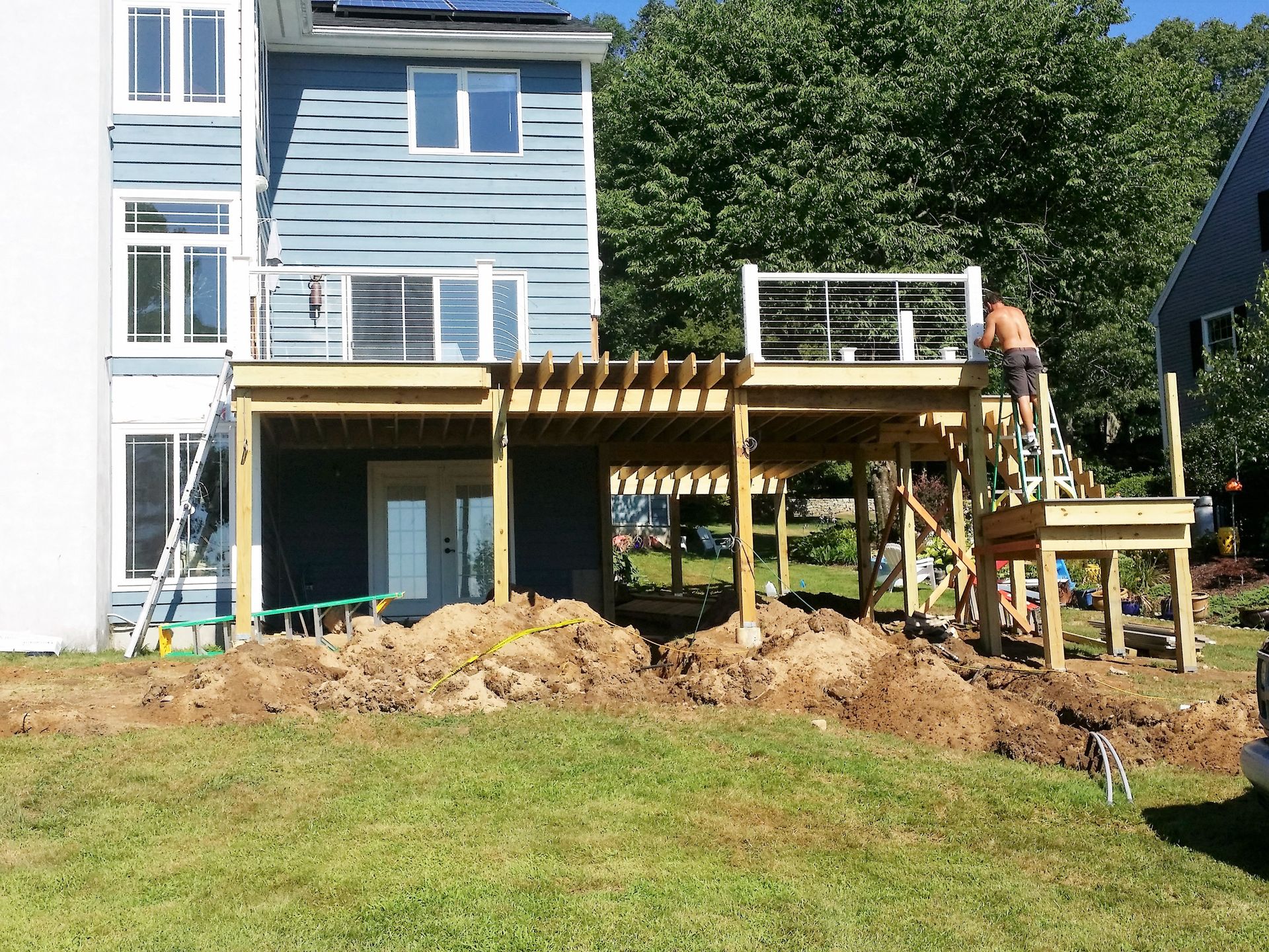 Construction of a multi-level wooden deck attached to a blue house in a grassy yard; worker on the deck.