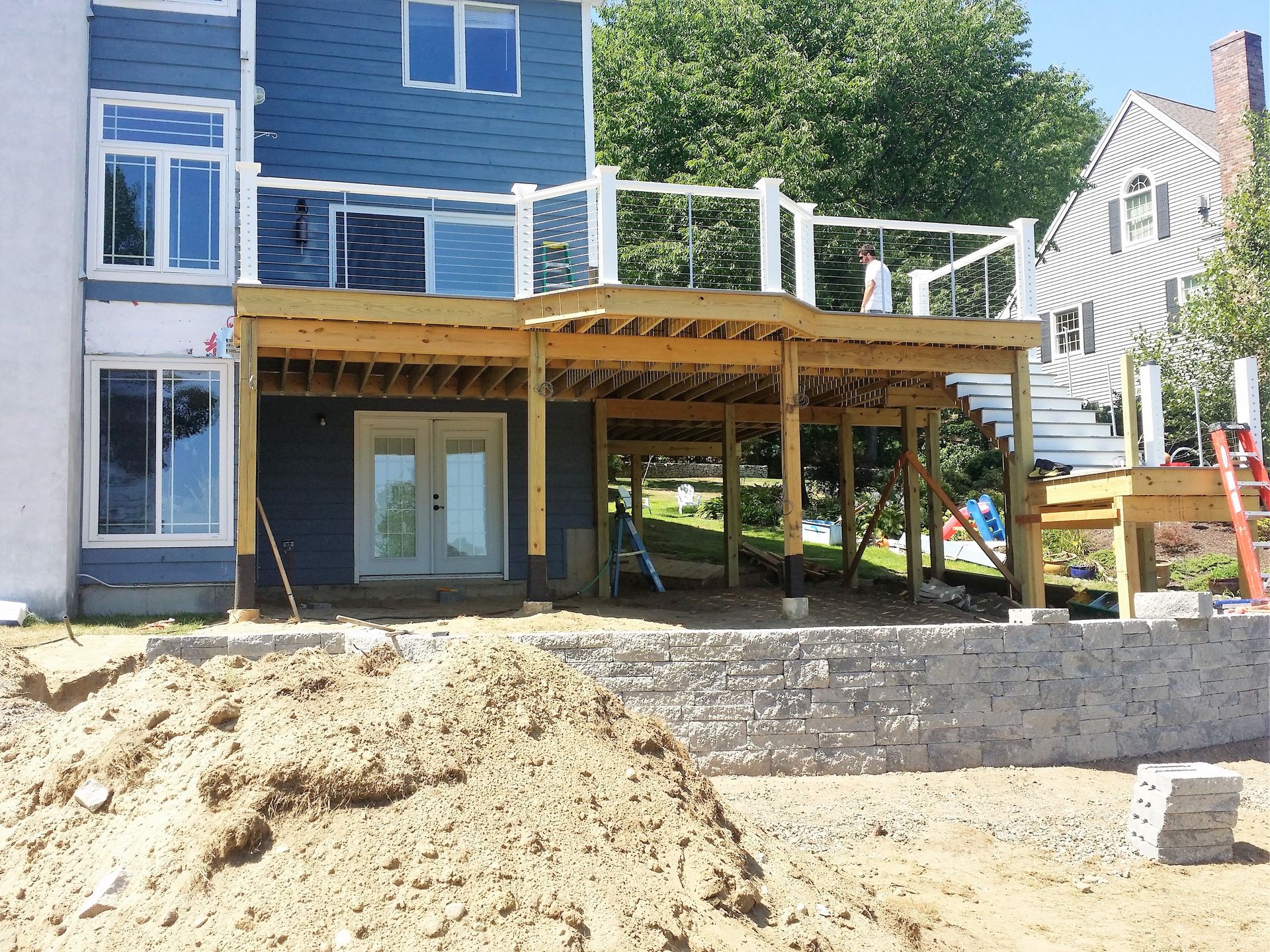 Deck construction in progress on a multi-story house with blue siding and a retaining wall.