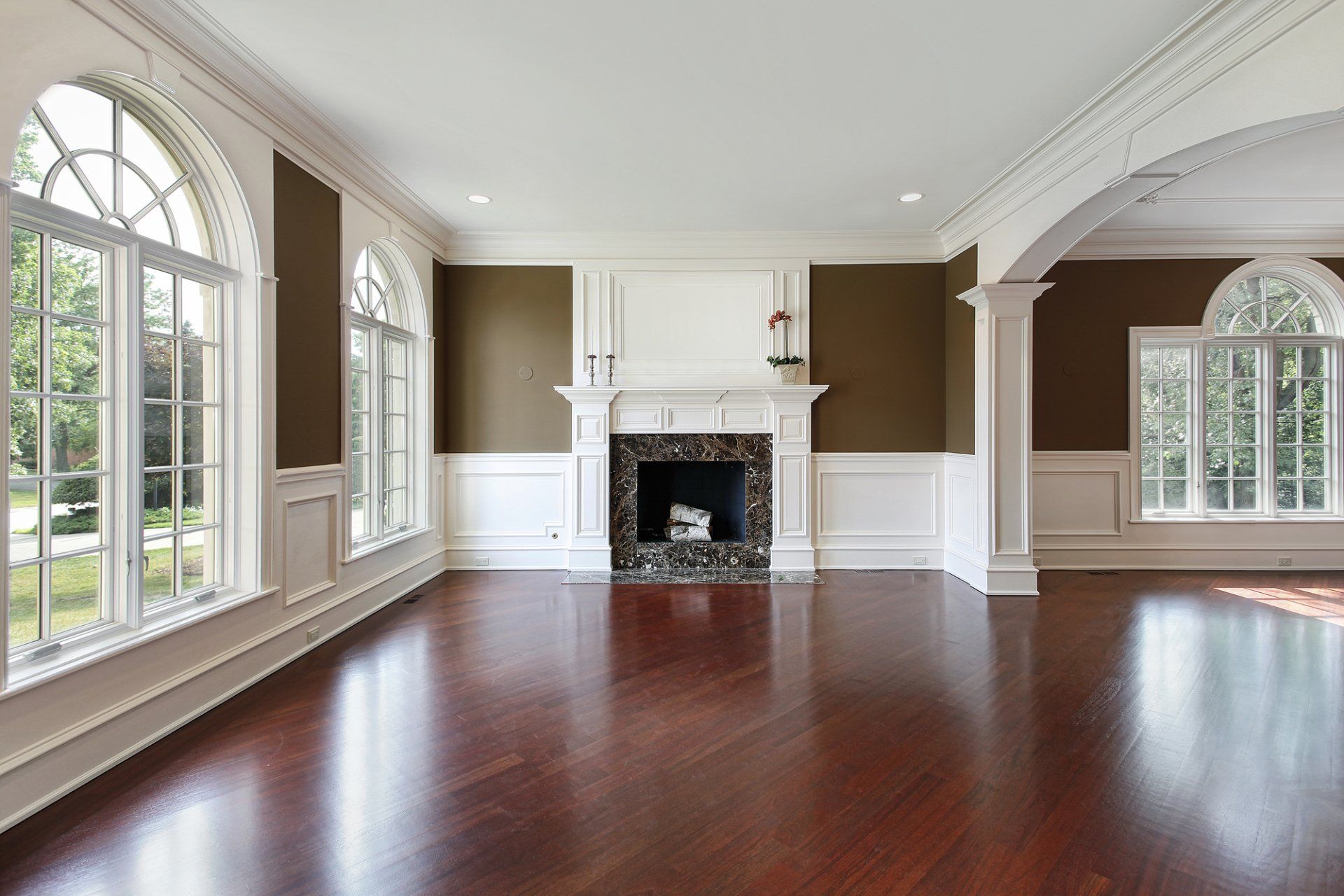 Empty living room with dark brown hardwood floors, white trim, fireplace, and arched windows.