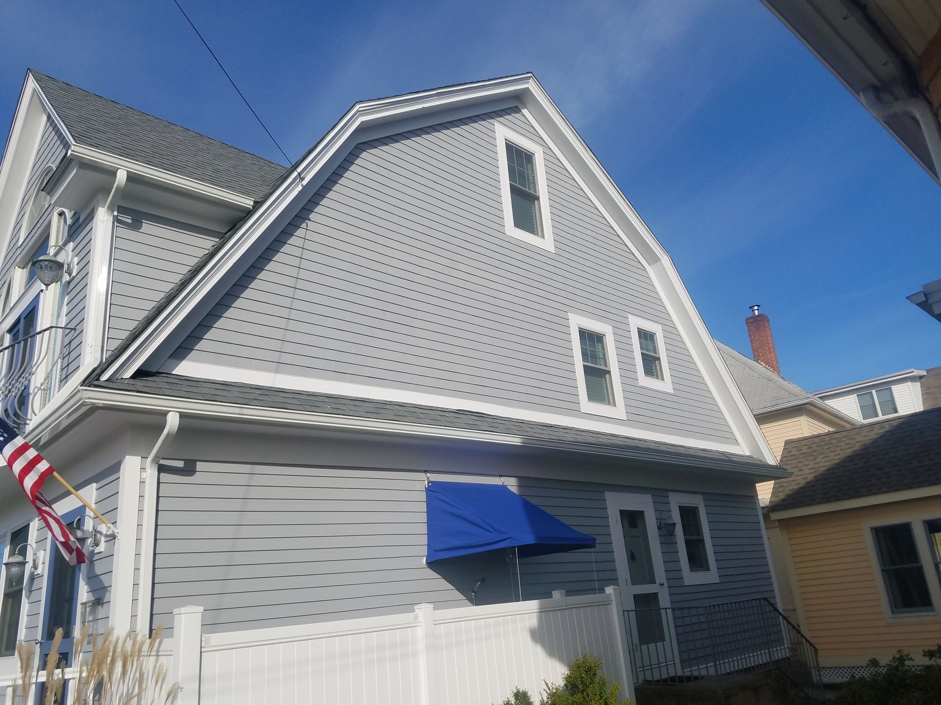 Gray house with white trim, blue awning, American flag, against a clear blue sky.