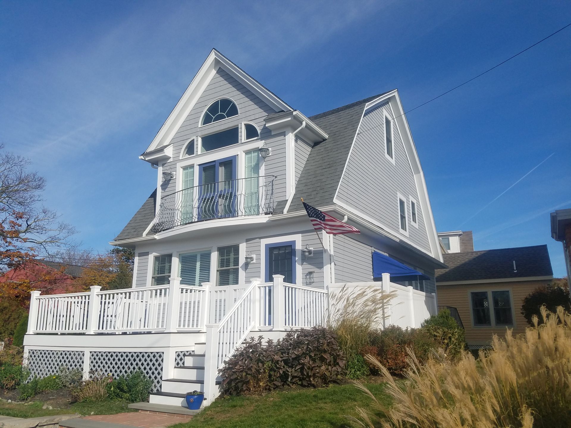 Two-story gray house with white trim, blue door, and a front porch, set against a bright blue sky.