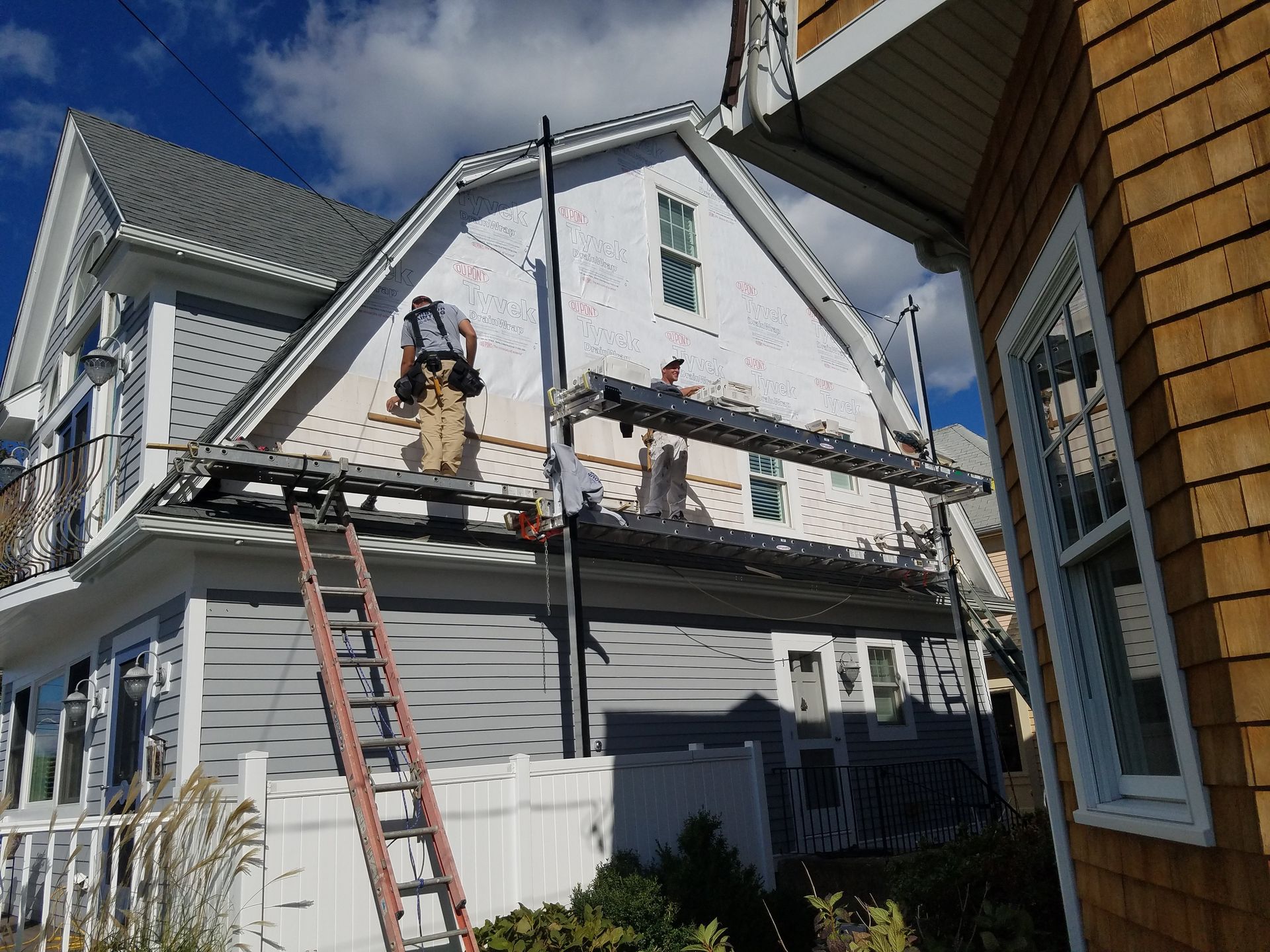 Workers on scaffolding installing siding on a two-story white house. Ladder in front, blue sky above.