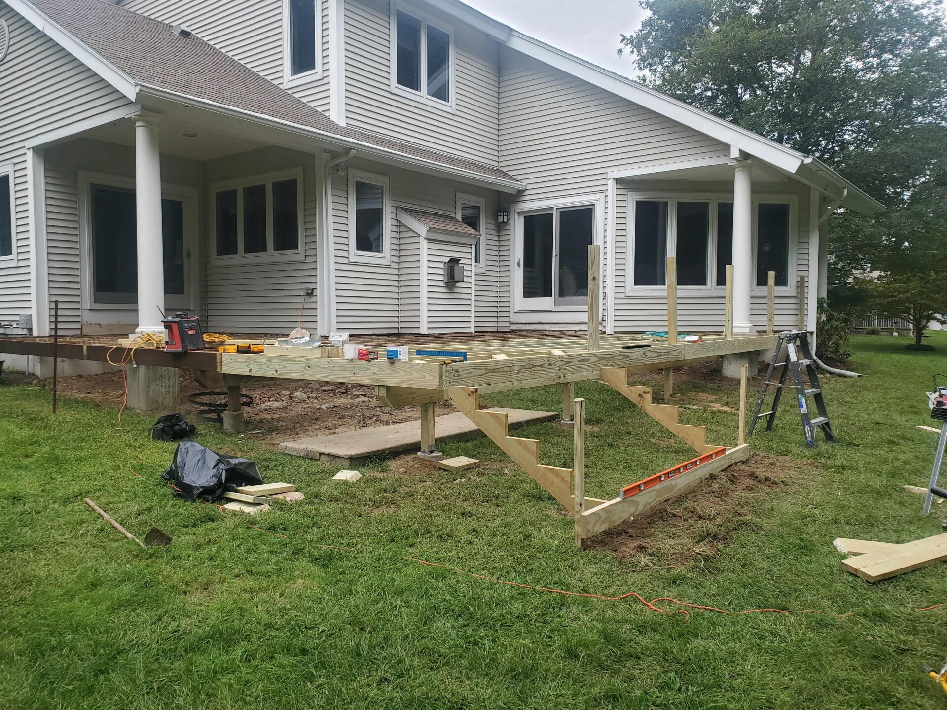 Deck construction in progress: wood framing, stairs, tools, house with white siding, green grass.