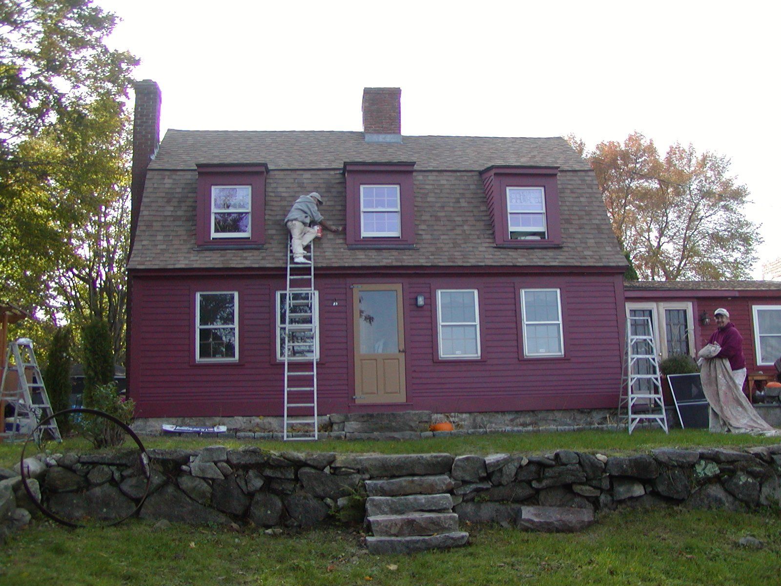 Red house with person on ladder, painting a window. Stone wall in front.