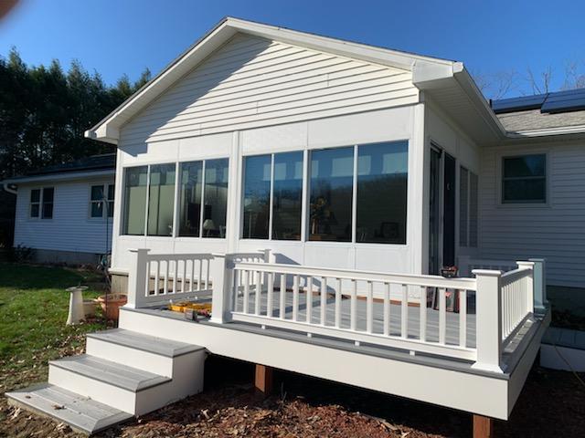 White sunroom with deck, gray steps and railing; attached to a house on a sunny day.