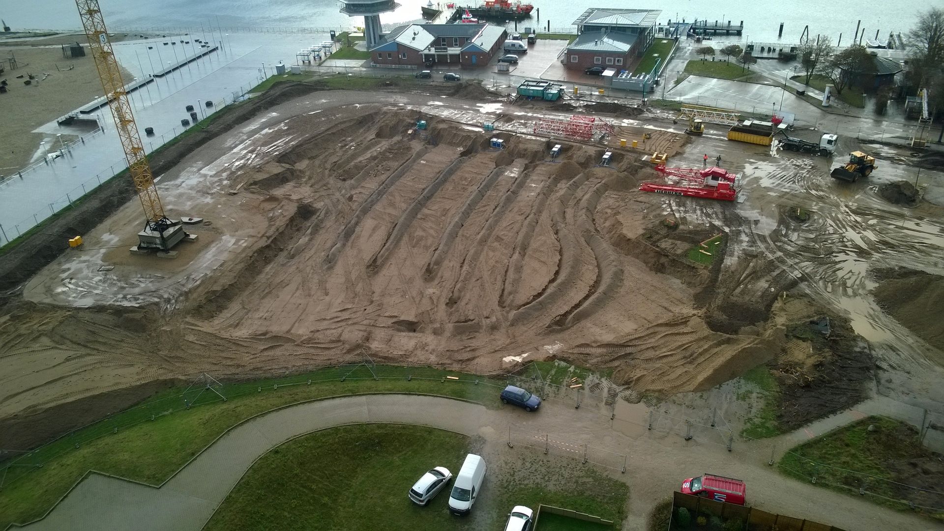 Baustelle am Wasser, mit schwerem Gerät und einer teilweise ausgehobenen Baugrube. Bewölkter Himmel.