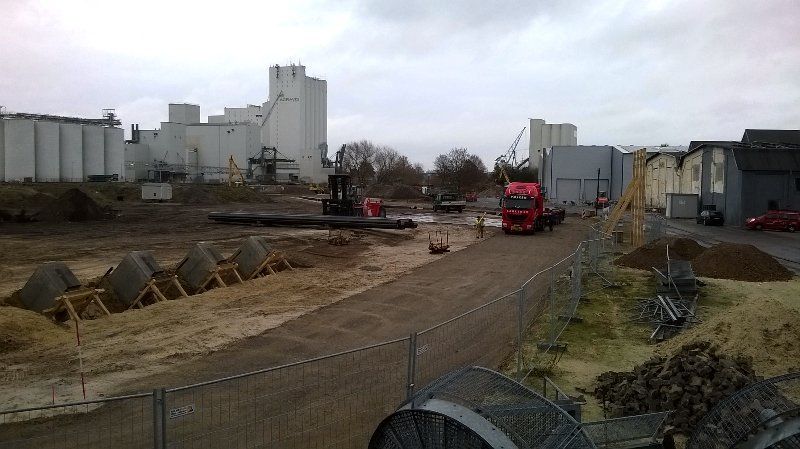 Baustelle mit großen weißen Industriegebäuden, schweren Maschinen und einem roten Lastwagen. Bewölkter Himmel.