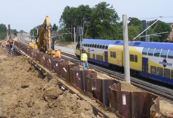 Baustelle entlang der Bahngleise: Bagger, Stahlbarrieren, vorbeifahrender Zug, anwesende Arbeiter.
