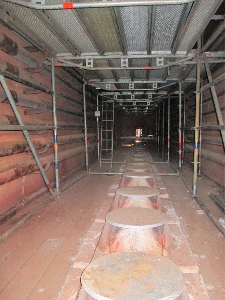 Innenansicht eines langen, schmalen Tunnels mit Gerüsten und großen, runden Metallgegenständen auf dem Boden.