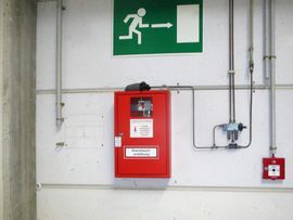 Roter Feuerlöschanlagenschrank an weißer Wand mit Notausgangsschild und Rohren.