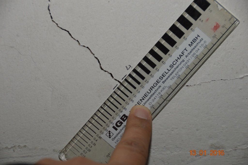 Eine Hand zeigt auf ein Lineal, um einen Riss in einer weißen Wand auszumessen.