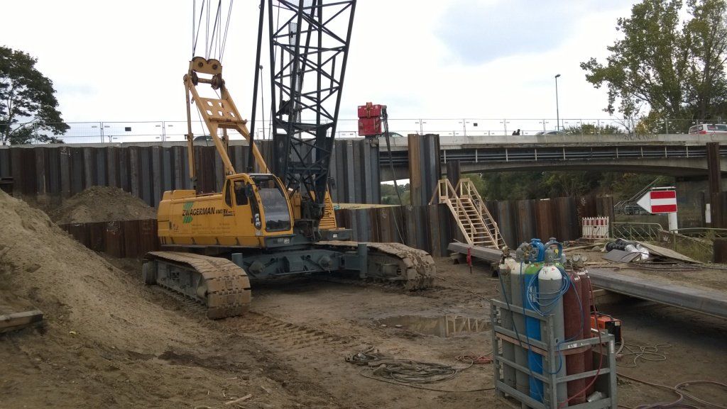 Ein gelber Kran auf einer Baustelle neben einer Metallwand und einer Brücke unter bewölktem Himmel.