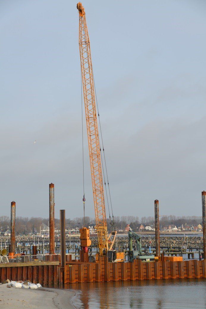 Baustelle mit orangefarbenem Kran, Strommasten und Wasser.
