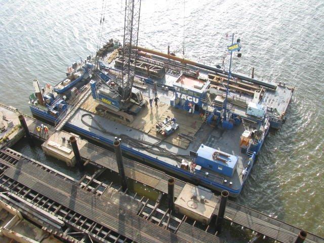Lastkahn mit Kran und Maschinen im Einsatz auf einer Wasserstraße. Grau und blau vor einem braunen Dock.