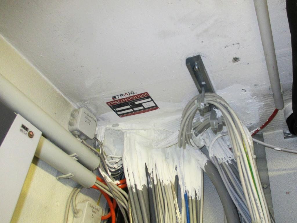 Zusammengebündelte Kabel und Drähte, mit weißem Dichtmittel überzogen, sind mit einer Metallhalterung an der Decke befestigt.