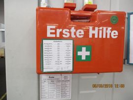 Ein orangefarbener Erste-Hilfe-Kasten mit der Aufschrift „Erste Hilfe“ und einem grünen Kreuz an der Wand.