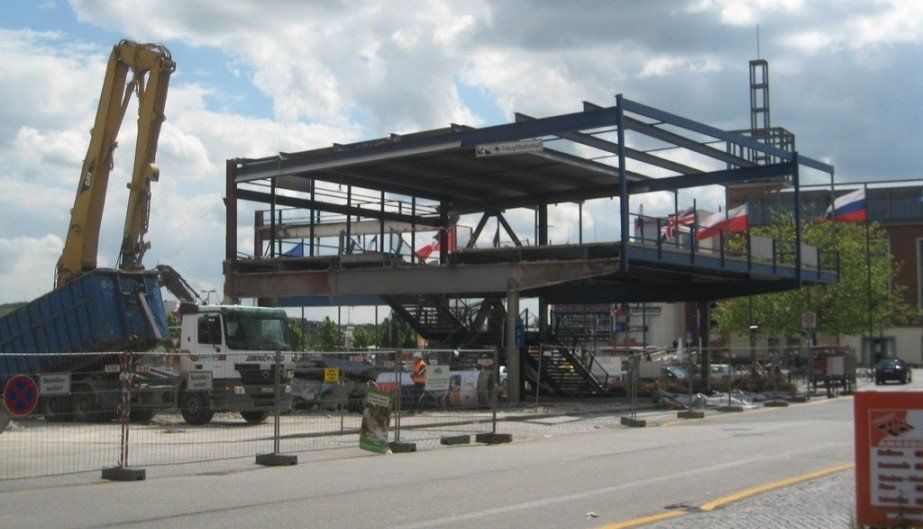 Abriss einer in der Luft hängenden Metallkonstruktion. Ein Kran und ein Lkw sind zu sehen, außerdem Trümmer und Bauzäune.
