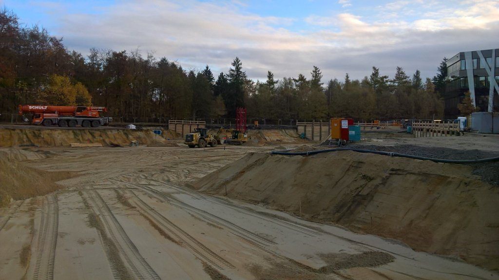 Baustelle mit ausgehobener Erde, schweren Maschinen und Bäumen im Hintergrund. Bewölkter Himmel.