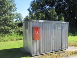 Verzinkter Metalllagercontainer mit rotem Feuermelderkasten an der Seite, auf einer Betonplatte im Freien aufgestellt.